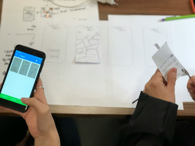 Person holding black iPhone with images of a design project on the screen. Another person cuts out paper for prototypes.

