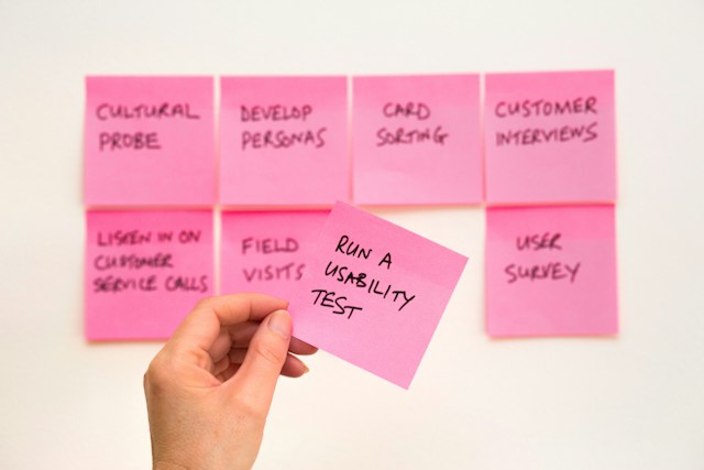 Two rows of eight pink post-it notes outlining the UX design process. A hand holds one reading "Run a Usability Test".