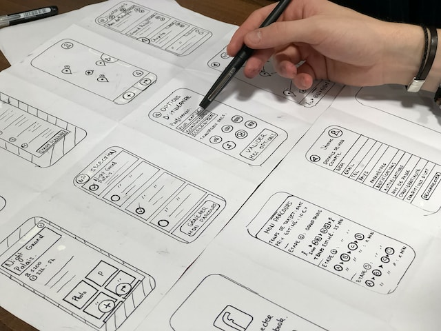 A person holds a pen near a piece of paper. The paper displays potential app layout designs.