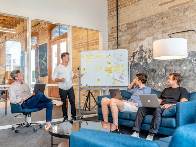 A product team makes notes on their laptops, surrounding a man beside a whiteboard. 