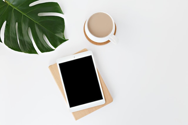 A white iPad on a white surface beside a cup of coffee and a plant.
