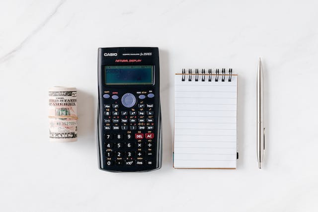 Flat lay image of a calculator next to a roll of paper money, a notebook, and a pen on a marble slab.