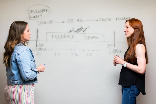 Two women talk beside a whiteboard with text written on it.