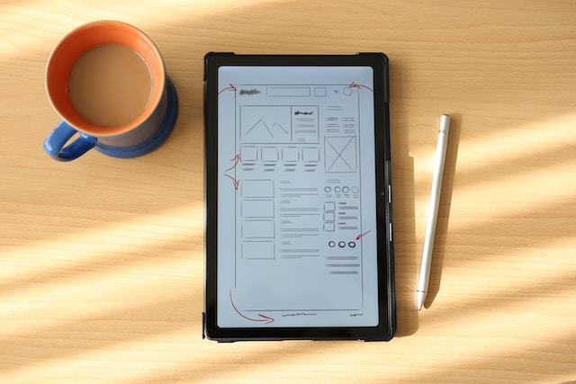 A tablet showing low-fidelity wireframes with red arrows for notes on a desk next to a stylus and cup of coffee.