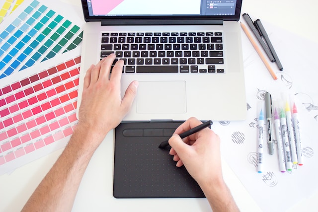 A designer works at a computer on a desk beside color charts and pens. 