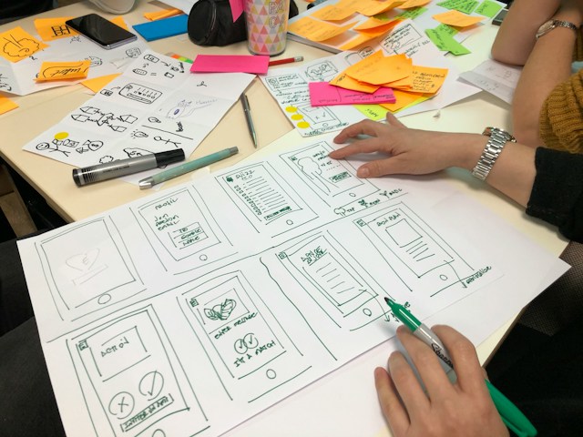 A person surrounded by sticky notes writes on white paper. The paper displays a series of web pages for a digital product. 