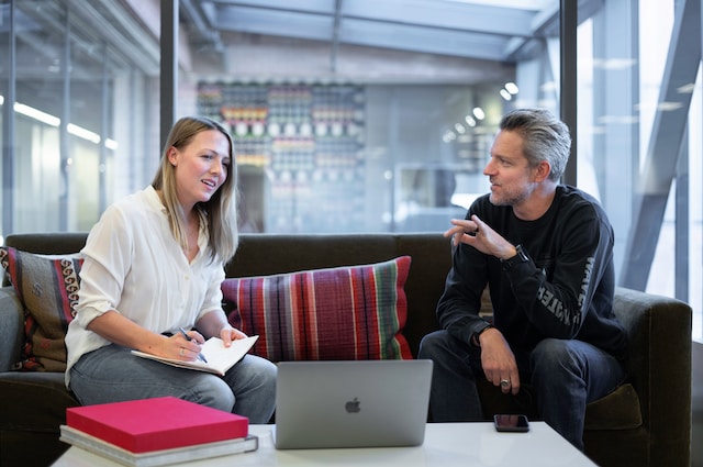 A man and a woman on a couch in a business office reference a laptop sitting on a table during their interview discussion.