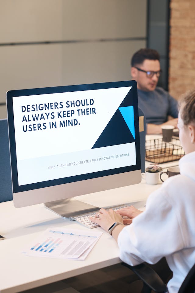 A person sits at a desk by a computer. The screen displays the words ‘Designers should always keep their users in mind.’ 