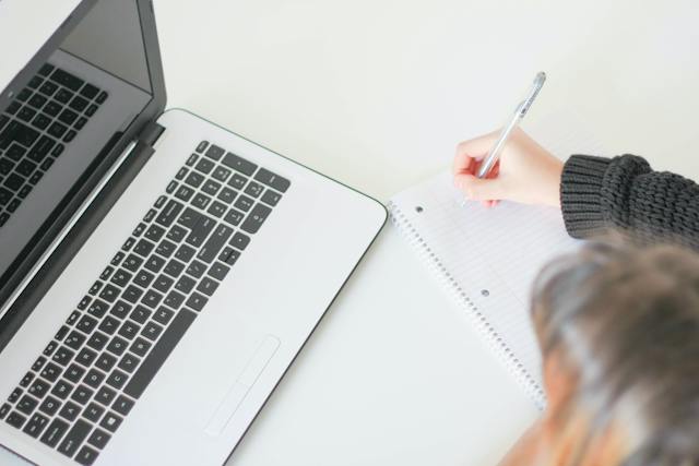 A woman writes in an empty notebook on a table. A turned-off MacBook is beside her on the table.