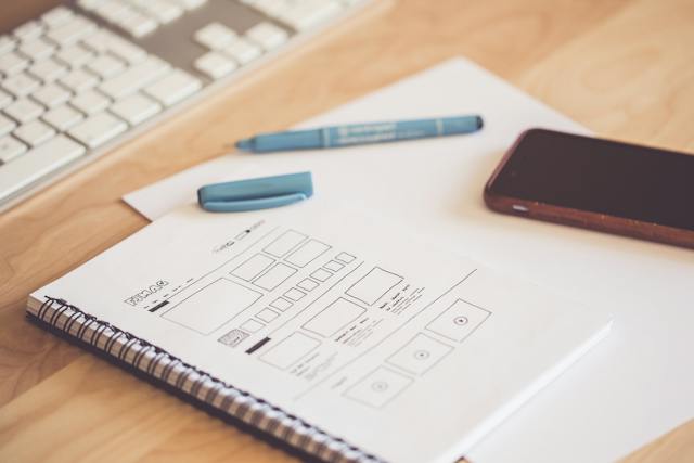 A blue pen with its lid off rests next to an open notebook with a wireframe drawn on it.