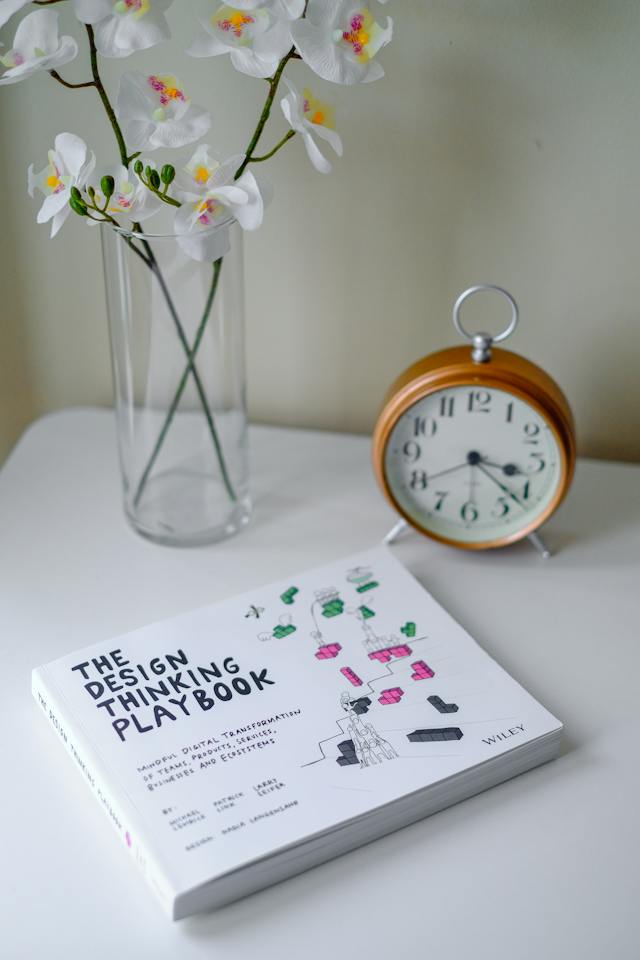 A book of the title ‘The Design Thinking Playbook’ sits on a white table near a clock and flowers. 