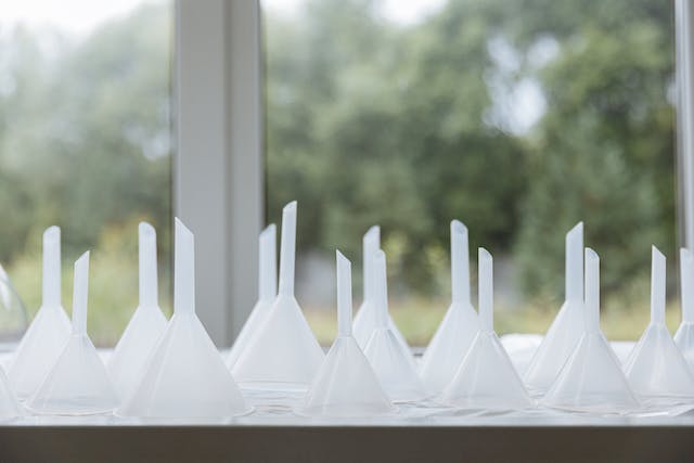 Many white plastic funnels lay with the spouts facing up on a counter in front of a window.