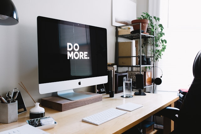 A turned-on iMac on a wooden desk, with a black screen reading "Do More" in capital letters.
