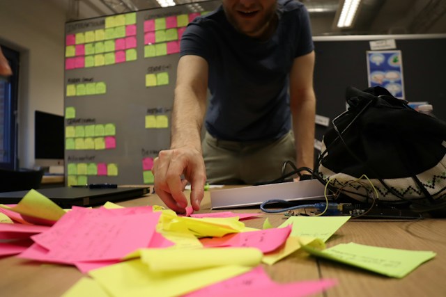 A man reaches for a pile of sticky notes on a table in front of him. Behind him is an affinity diagram.
