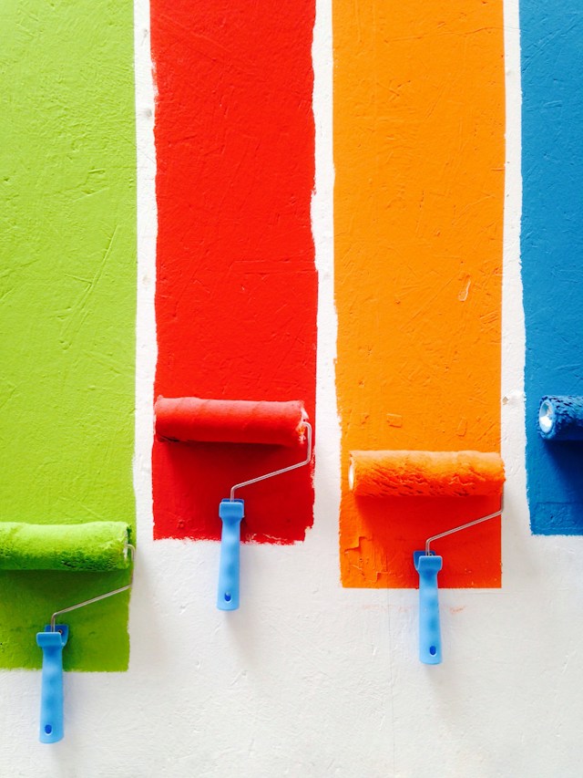 Four paint rollers appear pressed against a white wall. They paint orange, green, red, and blue colors on the wall. 