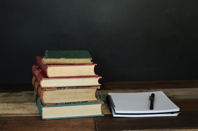 A stack of books sits next to a lined notebook. A black ballpoint pen sits on top of the notebook. 