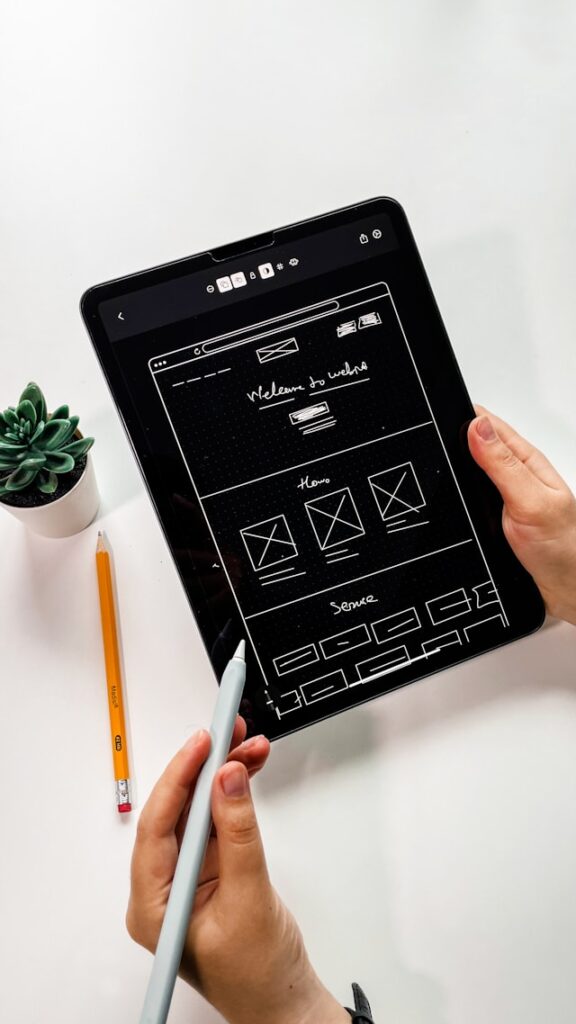 A person holds a black tablet with a wireframe sketched on the screen.

