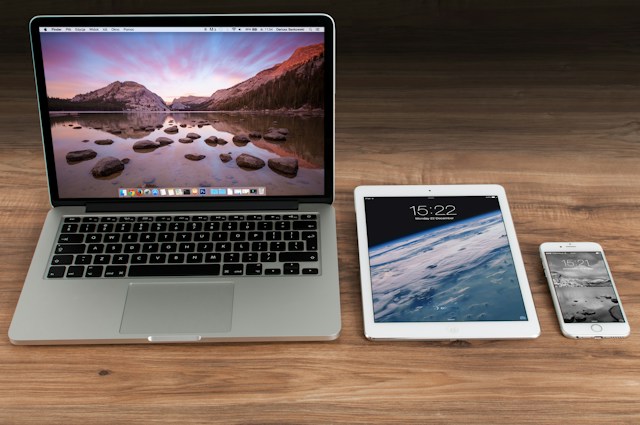 A turned-on Apple Macbook on a wooden table beside a turned-on iPad and a turned-on iPhone.