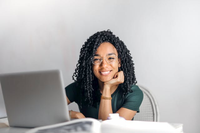 A smiling woman sits at a desk in front of an open laptop.
