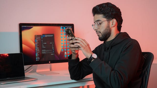 A man sits at a desk using his phone, with a turned-on desktop monitor in the background.
