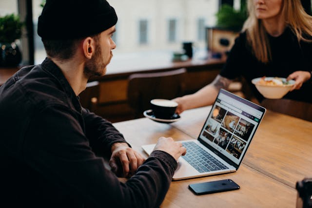 A man uses a turned-on laptop in a coffee shop as a waitress hands him coffee.
