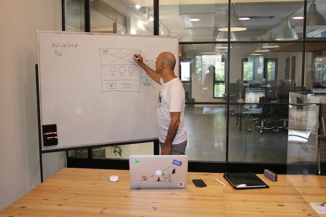 A man draws wireframes on a whiteboard. An open laptop is on the desk behind him.