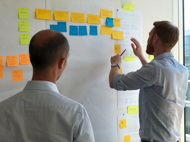 Two men look at a whiteboard covered in sticky notes. The two men collaborate on the design process of a product. 
