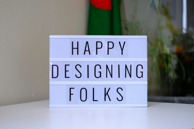 A light box sits on a white table. The phrase within the light box reads, ‘Happy designing, folks.’ 
