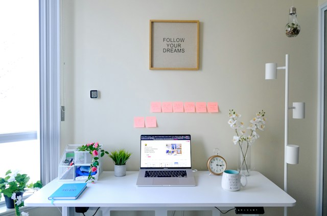 A turned-on laptop on a white desk with sticky notes taped to the wall in the background.