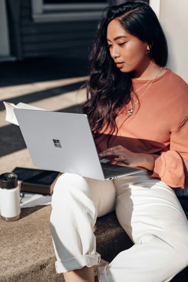 A woman sits on a surface outdoors using a Windows laptop.
