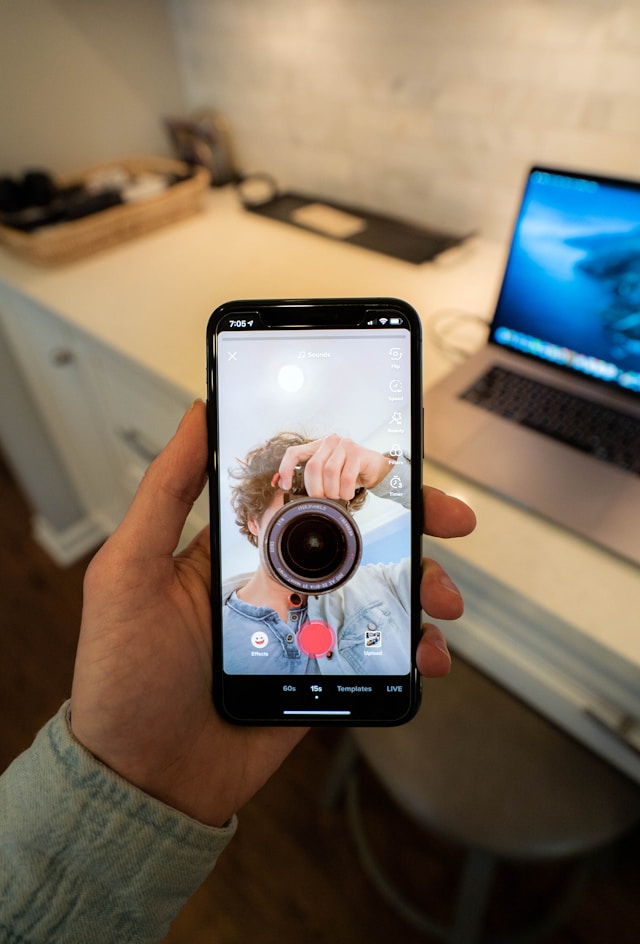 A person holds a turned-on smartphone open on the camera app. On the screen, a man holds a camera in front of his face.