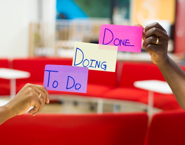 Two people hold three sticky notes arranged in line with words written 'To Do,' 'Doing,' and 'Done,' respectively.