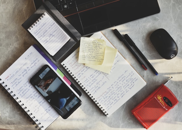 A desk with various things spread over it, including several notebooks, pens, and a turned-on phone.