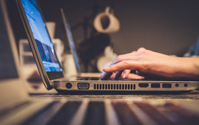 A side-on view of a turned-on laptop on a table, with two hands typing on the keyboard.