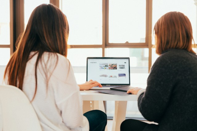 Two women talk while using a turned-on laptop on a white table.