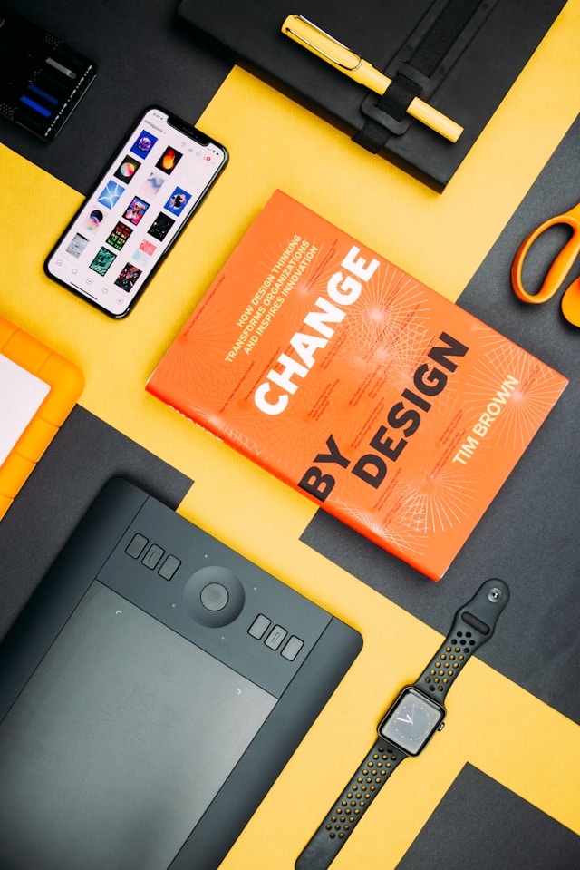 Various objects on a yellow table, including electronic devices and the “Change By Design” book by Tim Brown.