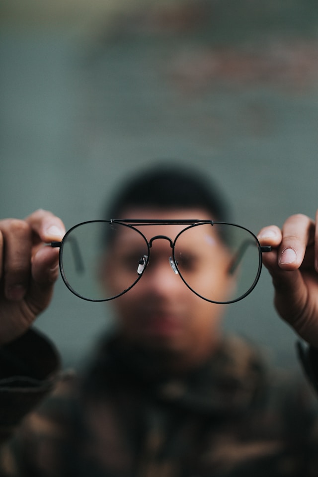 A person in an obscured background holds up their glasses to the camera. 
