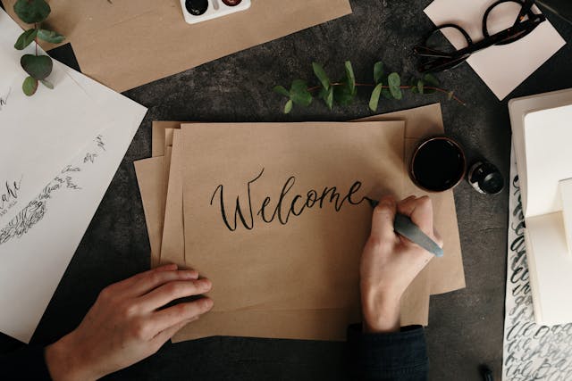 A person writes the phrase ‘Welcome’ in a sophisticated font on brown paper.