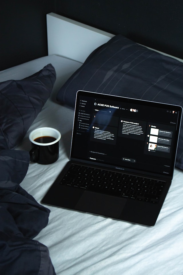 A black laptop sits on a white bed. The laptop’s screen displays the use of dark mode UI. 