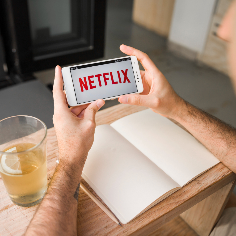 A person holds their white smartphone over a table. The phone’s screen displays the Netflix logo.

