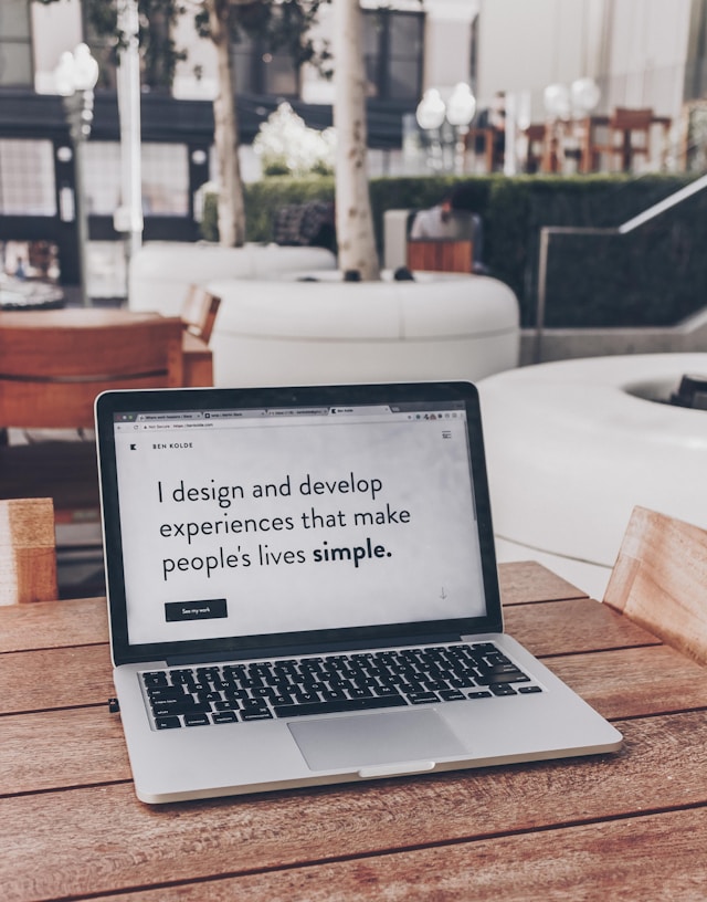 A laptop on a wooden table. The screen reads, "I design and develop experiences that make people's lives simple."
