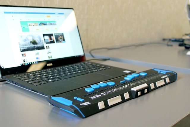 A laptop computer on a desk with a refreshable braille display attached.
