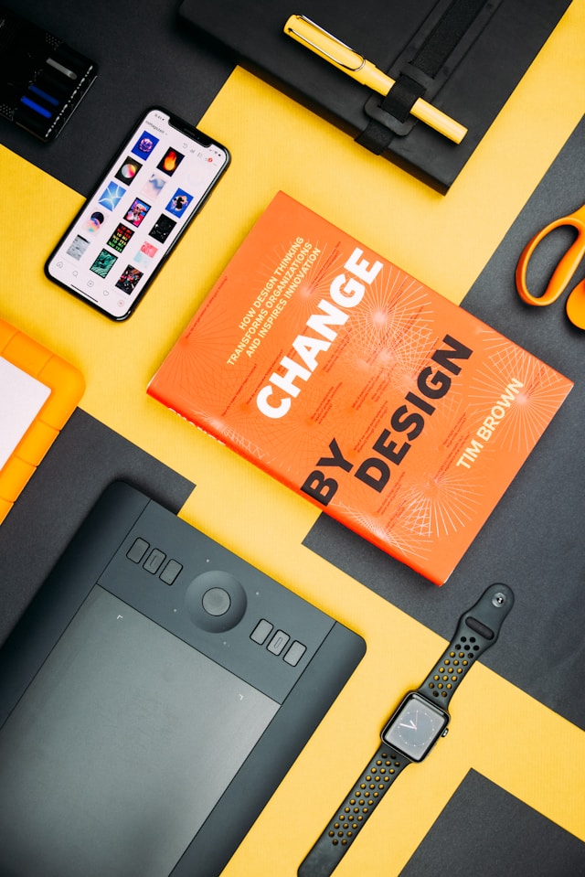 A collection of objects on a yellow background, including a Change By Design book, smartphone, and smartwatch.

