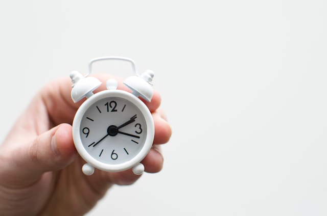A person holds a small white alarm clock, small enough to fit in just two fingers.
