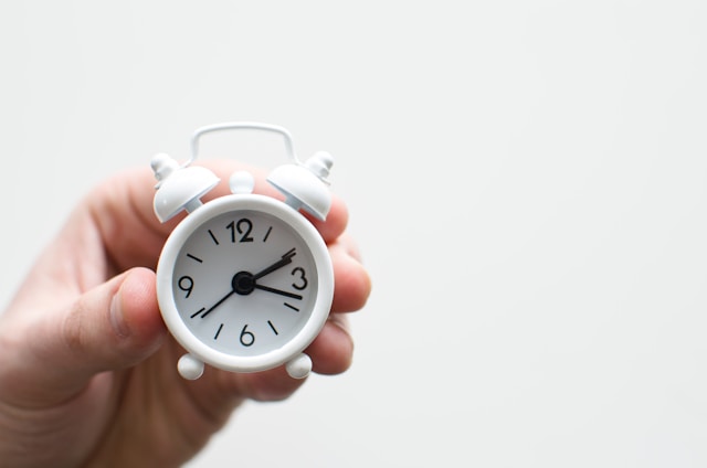 A person holds a small white alarm clock, small enough to fit in just two fingers.