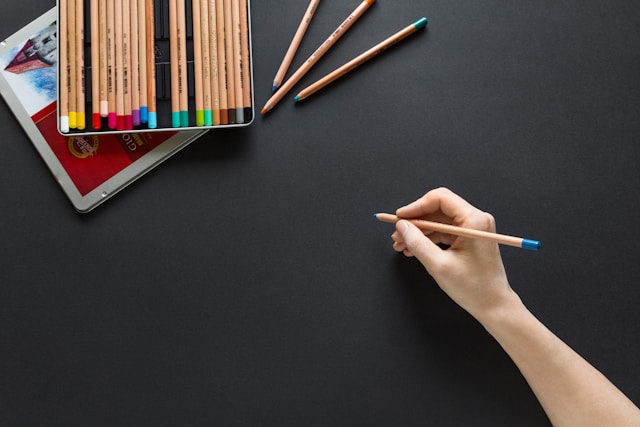 A person holds a blue pencil over black paper. A tray of colored pencils is at the top of the image.
