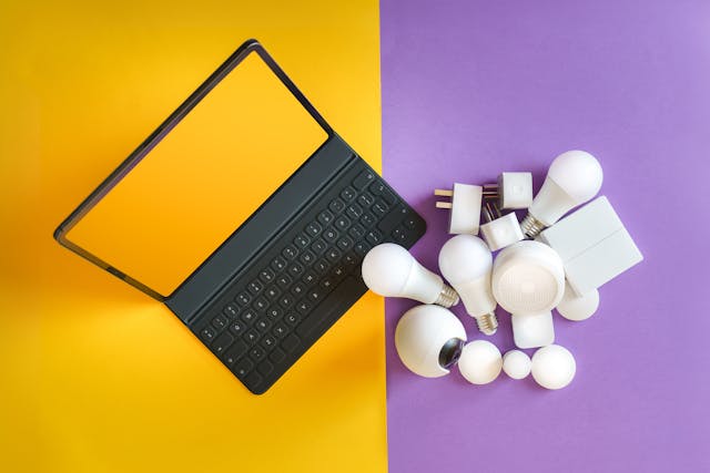 A laptop on a yellow and purple background with a collection of electronic items, such as plugs and bulbs.
