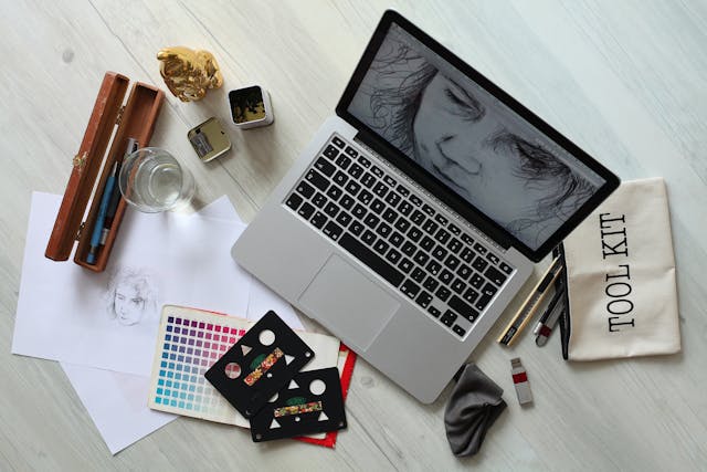 A grey laptop displays a digital illustration. Color swatches and art equipment surround the laptop. 
