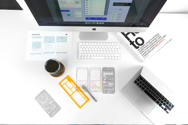 A shot from above of a white desk with numerous things on it, including a Mac, a laptop, paper wireframes, and a book.
