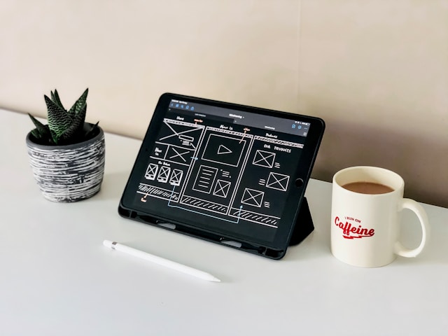 A black iPad sits beside a white mug on a white table. The screen displays a wireframe. 